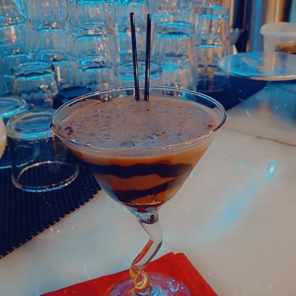 A chocolate martini with two straws in a twisted-stem glass, with stacked glasses in the background.